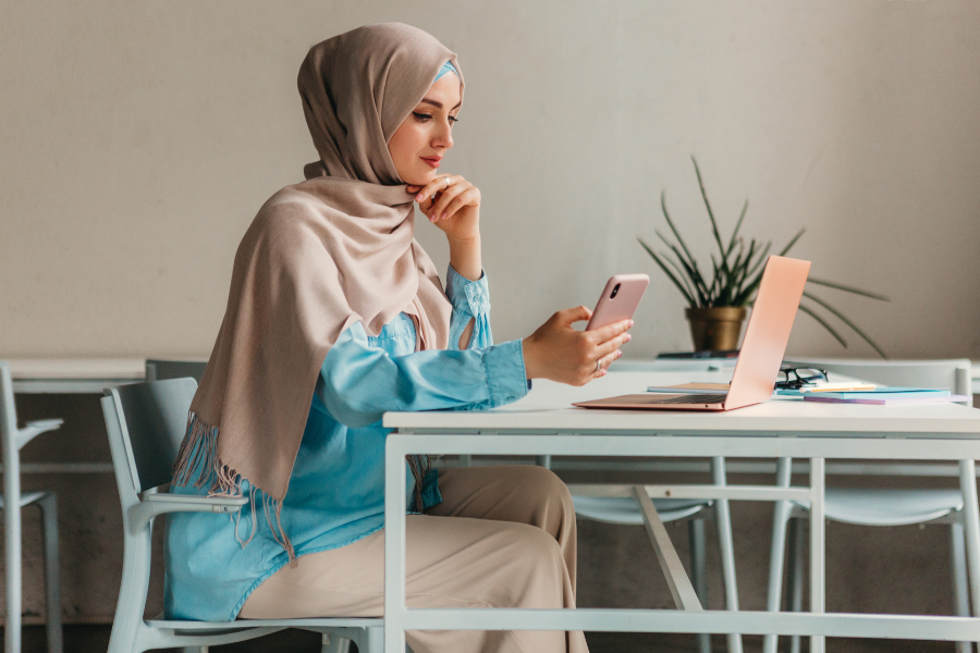 Businesswoman wearing a hijab using a smartphone beside her laptop in an office