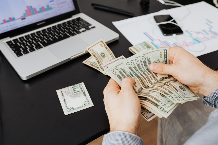 Person counting US dollar bills at a desk with a laptop and financial analysis